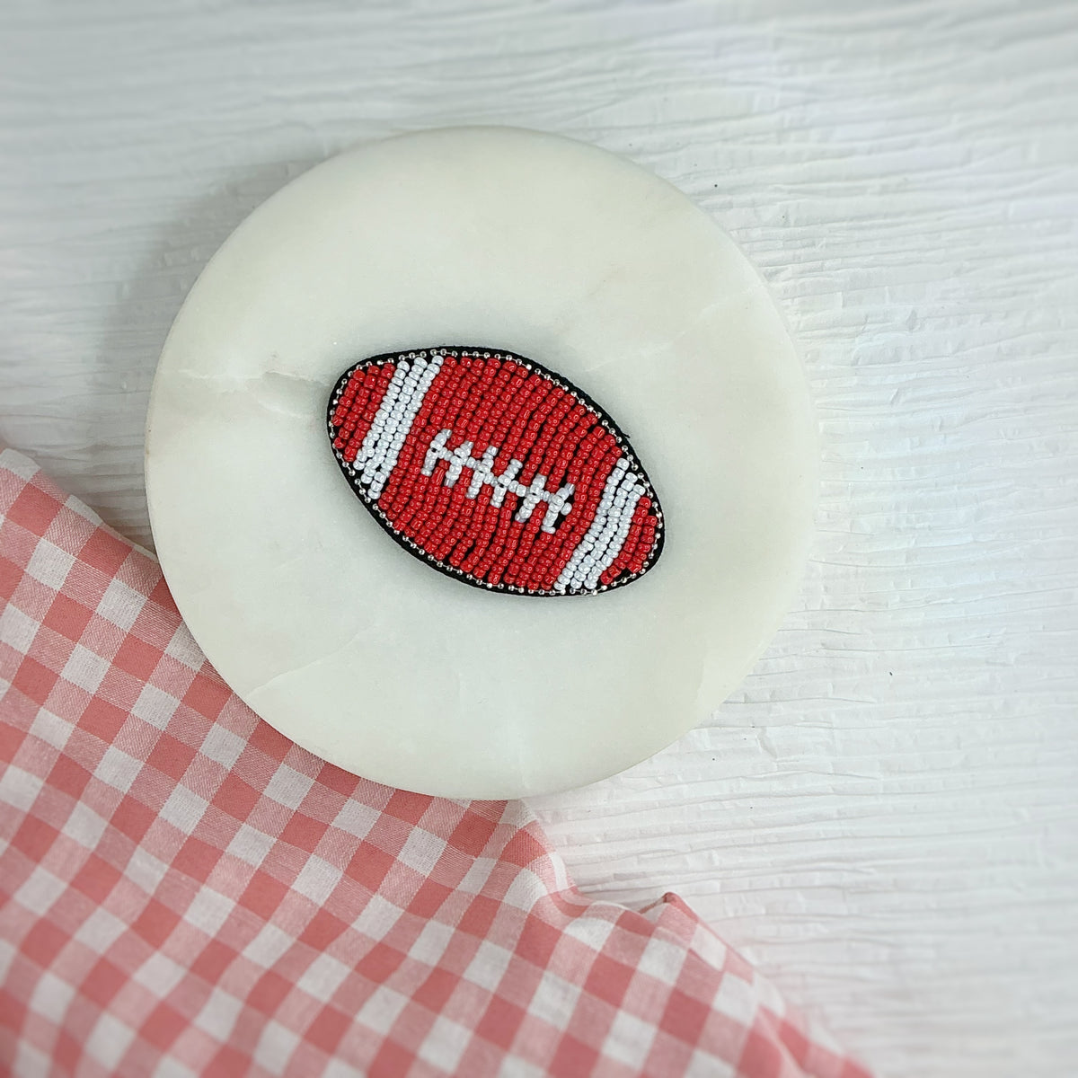 A round white dish featuring a central embroidered football patch in red, white, and black, styled on a white wood background with a pink and white checkered cloth in the corner.