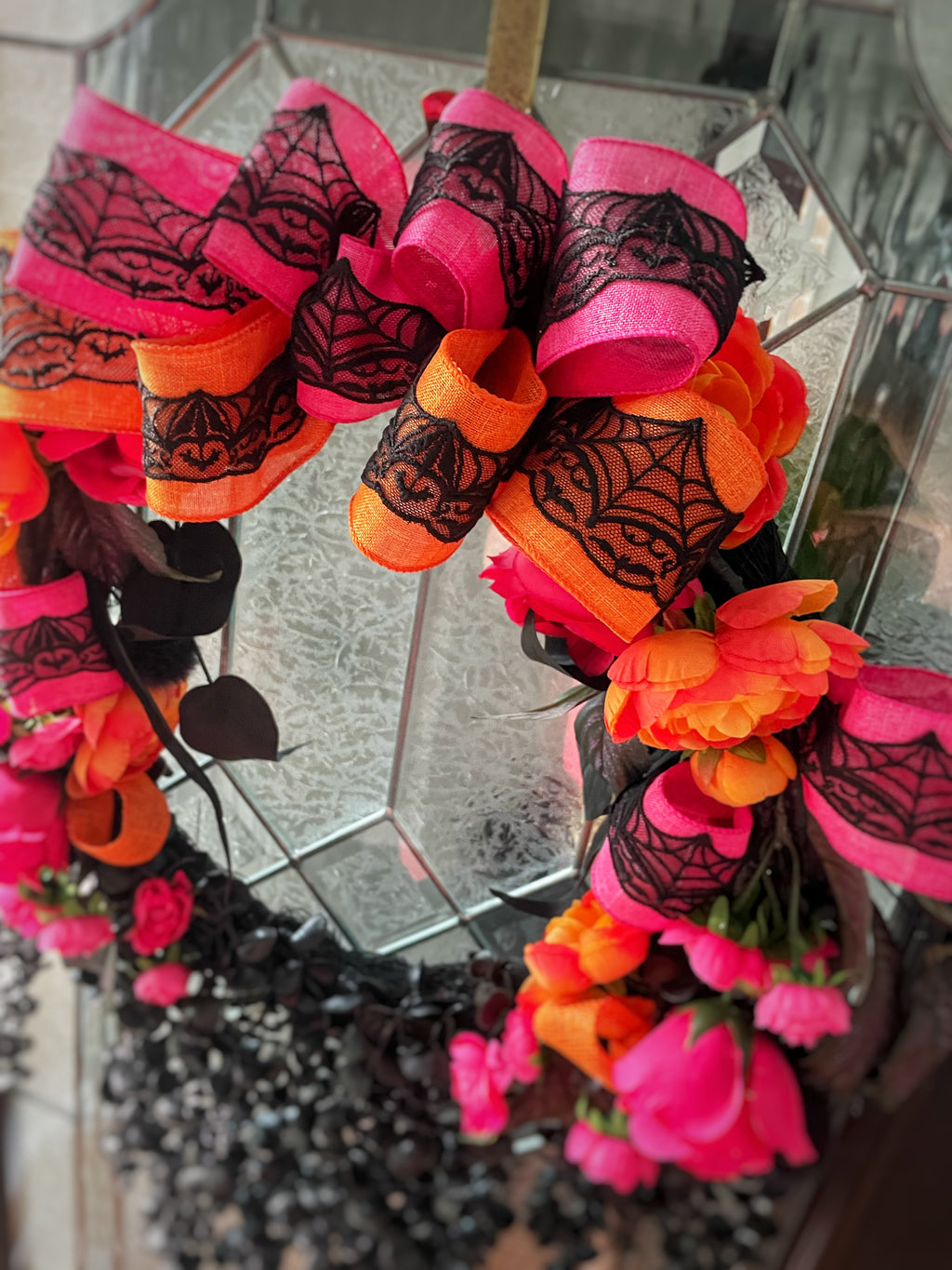 A close-up of a Halloween wreath decorated with bright pink and orange fabric flowers, black lace spiderweb patterns, and black ribbon accents, hanging in front of a mirrored glass surface.